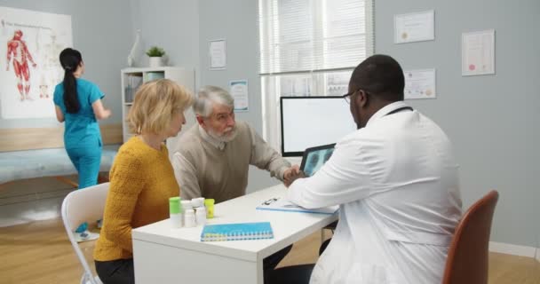 Specialista afroamericano di sesso maschile che tocca tablet discutendo e spiegando la scansione a raggi X per i pazienti anziani uomo e donna. Vecchia coppia caucasica in ambulatorio parlare con il medico concetto di salute — Video Stock