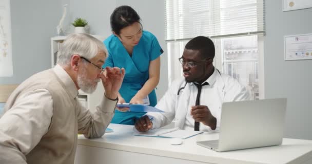 Porträt eines afroamerikanischen Arztes, der im Krankenhaus sitzt und mit einem älteren kaukasischen Patienten über Krankheiten spricht, während ihm eine asiatische Krankenschwester Dokumente aushändigt. Klinikkonzept — Stockvideo