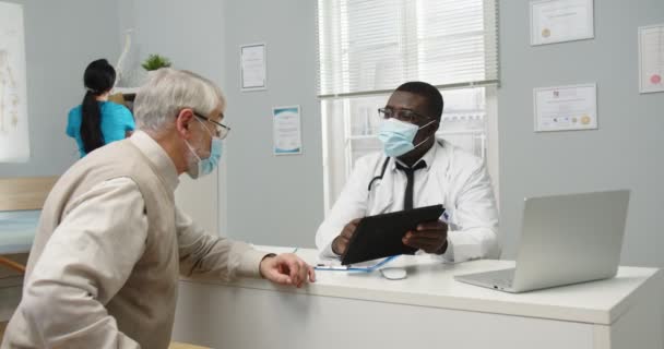 Ritratto di operaio ospedaliero afroamericano in maschera medica seduto in clinica e che parla con il paziente anziano caucasico mentre tocca con tablet. Malattia e malattia. Concetto medico — Video Stock