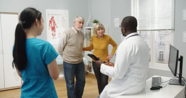 Parte trasera de afroamericano especialista médico hablando consultando a la vieja pareja de ancianos caucásicos acerca de resultados de la prueba tocando en la tableta. Pacientes felices en consulta de pie en la habitación del hospital — Vídeos de Stock