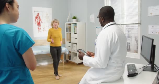 白人の高齢女性患者は、医師と一緒に診療室に立つ健康上の問題について話しますアフリカ系アメリカ人男性医師のリアタブレットを使用して病気についてシニア女性医師のコンサルティング病院の概念 — ストック動画