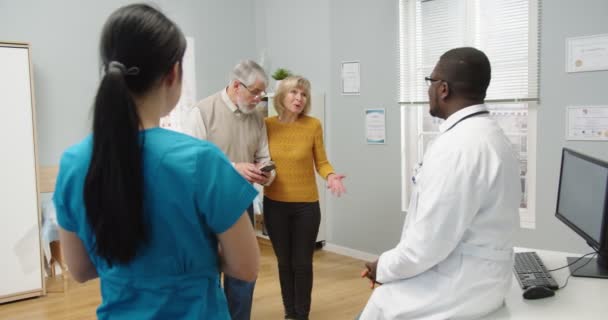 Parte trasera del médico afroamericano en consulta hablando consultando a viejos pacientes caucásicos de pareja de hombres y mujeres sobre la enfermedad o cirugía mientras están de pie en el gabinete del hospital. Concepto de salud — Vídeos de Stock