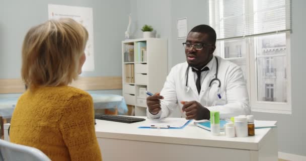 Retrato de un médico general afroamericano hablando y explicando problemas de salud y tratamiento a una paciente caucásica en consulta en el consultorio del hospital. Concepto sanitario — Vídeos de Stock