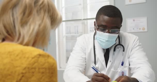 Närbild av snygg afroamerikansk manlig läkare specialist på mask sitter på jobbet i sjukhus chatta konsultera senior kaukasiska kvinna förskrivning och ge henne piller. Sjukvård, bakgrundsbild — Stockvideo