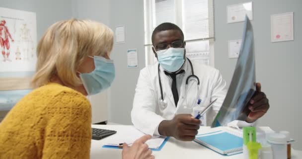 Primer plano del médico afroamericano en máscara médica sentado en el gabinete de la clínica, explicando y comentando la exploración por rayos X a una vieja paciente caucásica en consulta. Concepto de Coronavirus — Vídeos de Stock