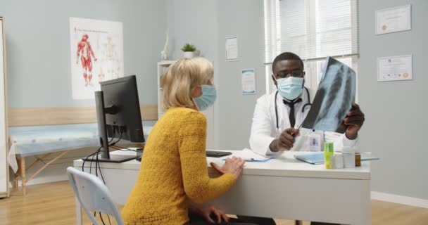 Homem afro-americano médico profissional em máscara médica na clínica sentado na mesa, falando com a velha mulher branca paciente em consulta explicando e comentando X Ray scan. Conceito de neurocirurgião — Vídeo de Stock