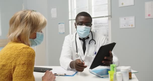 Primer plano del médico profesional afroamericano en máscara médica sentado en el gabinete de la clínica, explicando el resultado de la prueba de tratamiento a una vieja paciente caucásica usando tableta en consulta. — Vídeos de Stock