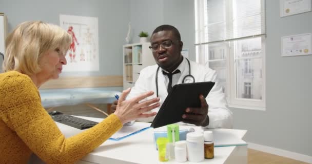 Zaměřil se na afroamerického lékaře v lékařském kabátě, který seděl u stolu a radil se se starší pacientkou o nemoci nebo chirurgickém zákroku, který ťukal na tablet. Detailní záběr ženy mluvit s lékařem na schůzce — Stock video