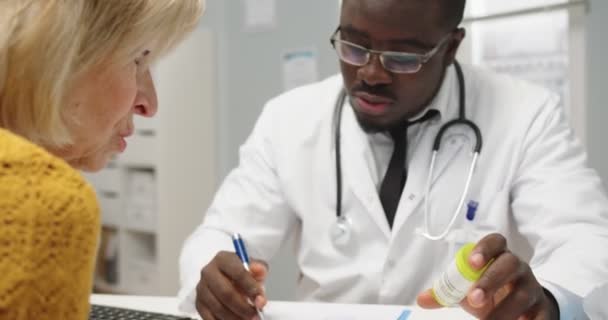 Großaufnahme eines gutaussehenden afroamerikanischen jungen Arztes mit Brille, der am Schreibtisch in der Klinik sitzt und Medikamente und Pillen für ältere kaukasische Frauen verschreibt. Gesundheitskonzept — Stockvideo