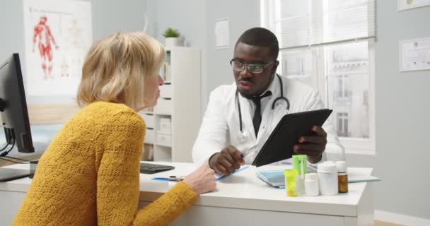 Retrato de un guapo médico afroamericano en gafas que explica la consulta de una paciente mayor caucásica usando tableta que muestra los resultados de la prueba. Parte trasera de la mujer en el hospital en consulta — Vídeos de Stock