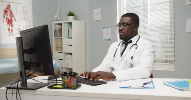 Portrait de l'homme professionnel afro-américain beau médecin en manteau médical dans des lunettes assis à table dans la chambre d'hôpital, travaillant sur la saisie informatique sur le lieu de travail et regardant la caméra Concept de clinique — Video