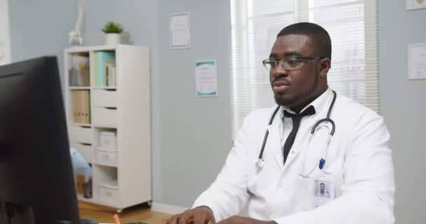 Portrait d'afro-américain occupé beau médecin professionnel masculin avec stéthoscope dans des lunettes assis au bureau dans l'armoire de l'hôpital, travaillant et tapant sur ordinateur au travail, regardant la caméra — Video