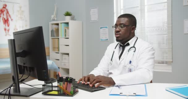 Portrait of serious African American busy professional male physician in glasses sitting at desk in hospital cabinet, working and tapping on computer, writing down information in clinical history — Stock Video