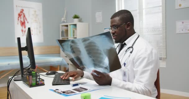 Портрет афроамериканского врача-мужчины в очках, сидящего за столом в больничном кабинете и печатающего на компьютере, глядя на результаты рентгеновского сканирования. Лечение заболеваний. Концепция клиники — стоковое видео