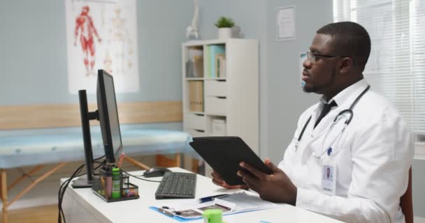 Retrato de un médico afroamericano serio enfocado en gafas sentadas en la mesa en el gabinete del hospital navegando y escribiendo en la tableta, mirando el monitor de la computadora. Tratamiento de enfermedades. Vista lateral — Vídeos de Stock