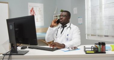 Ciddi derecede yakışıklı Afrikalı Amerikalı profesyonel bir doktorun portresi hastane kabinesinde oturmuş hastayla cep telefonuyla konuşuyor, telefonda danışmanlık yapıyor. Sağlık hizmetleri kavramı
