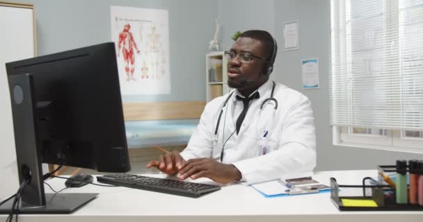 Occupé beau médecin professionnel afro-américain assis à la table dans l'armoire de l'hôpital parlant en vidéoconférence casque sur ordinateur, regardant moniteur. Concept de portrait. Soins de santé — Video