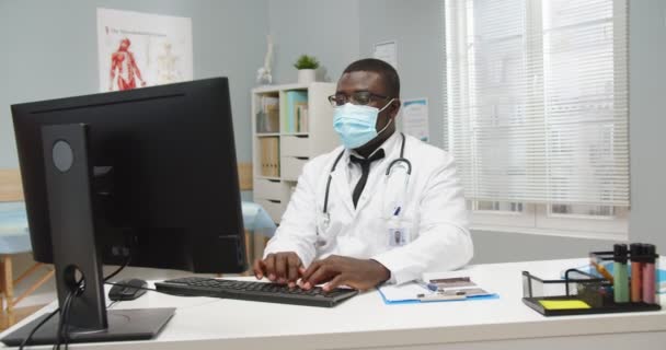 Ocupado bonito afro-americano masculino profissional médico especialista vestindo máscara médica sentado à mesa no armário do hospital mensagens de texto e digitação no computador, olhando para a câmera. Conceito de retrato — Vídeo de Stock