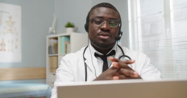 Primer plano del guapo médico profesional afroamericano ocupado sentado en el gabinete del hospital hablando en auriculares en video llamada en línea, videochat en computadora, concepto de atención médica — Vídeos de Stock