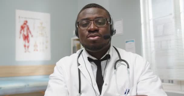 ビデオ電話でヘッドセットで話す病院のキャビネットに座っているハンサムなプロのアフリカ系アメリカ人男性医師のクローズアップ,ウェブカメラでビデオ撮影,医療.診療所相談のコンセプト — ストック動画
