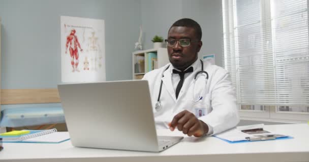 英俊而严肃的非洲裔美国全科医生的画像，坐在医疗中心的橱柜里，一边工作，一边在笔记本电脑上打字，一边搜索互联网。健康概念 — 图库视频影像