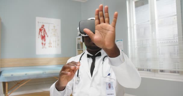 Крупный план афроамериканского врача-мужчины в белом медицинском халате, сидящего в больничном кабинете в очках VR, движущего руками в воздухе, работающего в виртуальной реальности. Инновационные технологии — стоковое видео