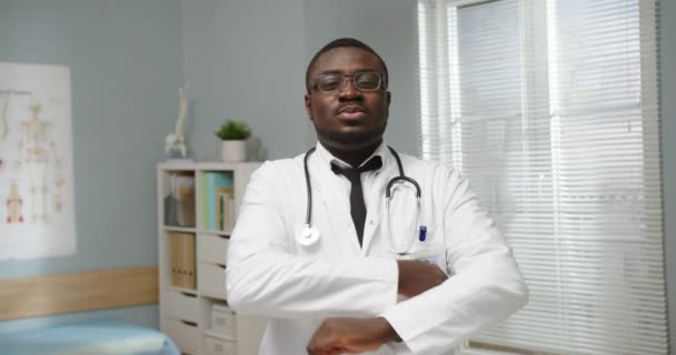 Retrato de bonito jovem afro-americano profissional de saúde com casaco branco e estetoscópio em pé no escritório do hospital sozinho, olhando para a câmera. Conceito de clínica — Vídeo de Stock