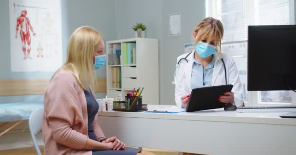 Medikal maskeli hoş sarışın bir kadının tablet bilgisayardan kadın doktorla test sonuçlarına bakarken orta boy fotoğrafı. Hastanın sağlık geçmişini incelemek için portatif tablet kullanan bir doktor.. — Stok video