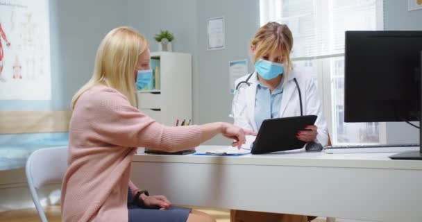 Colpo medio di bella giovane donna bionda in maschera medica guardando e indicando i risultati dei test su tablet con medico femminile. Medico che utilizza tablet portatile per esaminare la storia sanitaria del paziente. — Video Stock