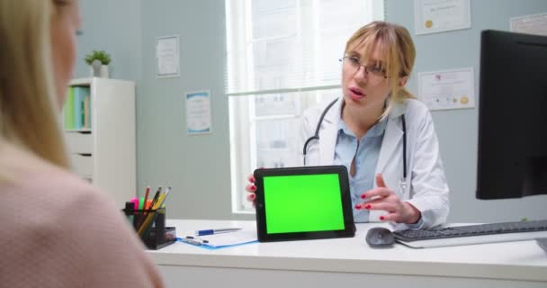 Sarışın güzel bir kadının, kadın doktorla tablet yeşil ekranda test sonuçlarına bakarken orta boy görüntüsü. Tıbbi doktor hastanın sağlık geçmişini incelemek için portatif tablet kromaanahtarı kullanıyor.. — Stok video