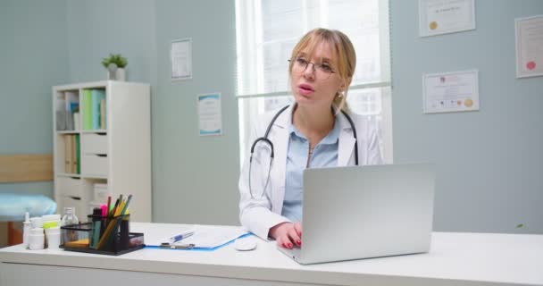 Primo piano del medico donna in occhiali guardando computer portatile. Donna bionda in visita medica. Specialista medico che fornisce servizi sanitari in ufficio ospedaliero. Sanità, concetto di medicina — Video Stock