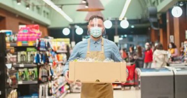 Handsome Caucasian man wearing face visor carrying box with vegetables in supermarket. Male grocery store owner in medical mask walking through products sections in store. Business, virus concept.