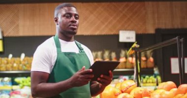 Attractive male worker making reports and checking products in grocery store. Young Afro-American guy in apron putting data on tablet in supermarket mall. Commerce, technology, marketing concept.
