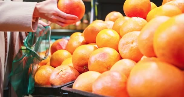 Nær kvinnelige hender som plukker appelsiner og putter i øko-pose i matbutikken. Ung kvinne som kjøper frisk moden frukt i supermarkedet for å øke immuniteten ved pandemi av coronavirus. Helse. – stockvideo