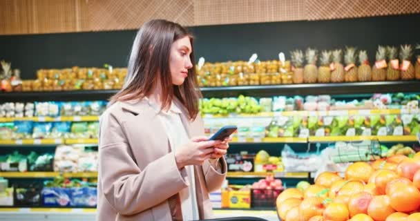 Erfolgreiche Kaukasierin checkt ihre Einkaufsliste im Supermarkt per Smartphone. Junge Frau mit digitalem Gerät und Blick auf Früchte im Supermarkt. Technik, Kaufkonzept. — Stockvideo
