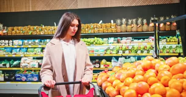 Mooie blanke vrouw die winkelwagentjes duwt en producten koopt in de supermarkt. Jonge vrouwelijke klant die fruit, sinaasappels en zet in eco-zak in supermarkt. Handel, bedrijfsconcept. — Stockvideo