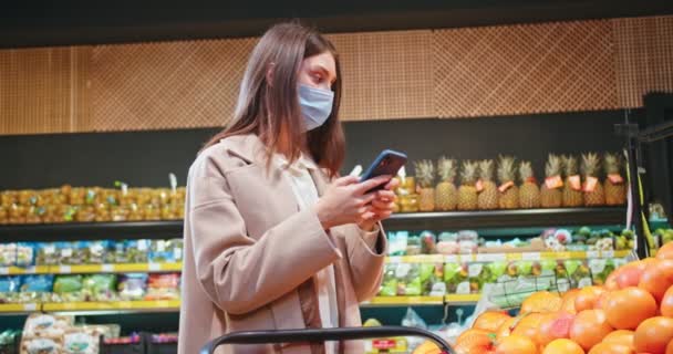 Beautiful Caucasian woman wearing medical mask and buying products in supermarket. Adult lady standing near fruit section and calling her mom to ask if she needs sth. Virus, safety, health, vitamins. — Stok video