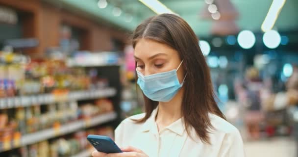 Schöne junge Frau in medizinischer Maske kauft Produkte im Lebensmittelgeschäft. Kaukasische Kundin checkt Einkaufsliste auf Smartphone im Supermarkt. Technologie, Einkauf, Covid-19-Konzept. — Stockvideo