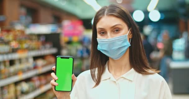 Attraente cliente donna che indossa mascherina medica che tiene smartphone con chiave cromatica. Giovane donna che mostra schermo verde e guardando la fotocamera nel supermercato. Quarantena, concetto di pandemia. — Video Stock