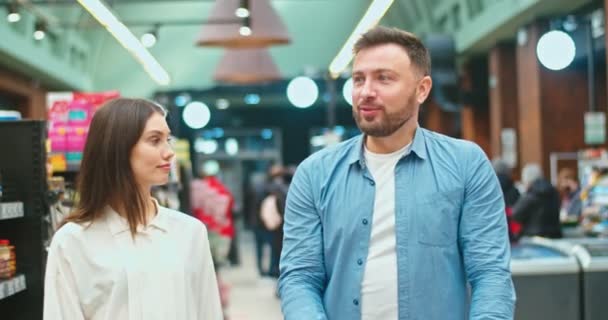 Sweet Caucasian couple buying ingredients to cook romantic dinner. Man and woman talking, pushing shopping cart and taking pickles in supermarket. Love, family, relationships, marriage concept. — Stok video