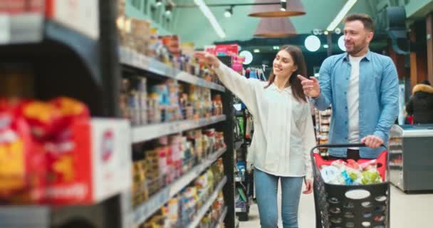 Sweet young family choosing products for cooking dinner together in supermarket. Caucasian couple with shopping cart buying groceries and talking to each other. Marriage, purchase concept. — Stok video