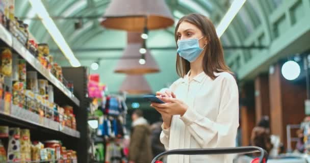 Una mujer guapa usando máscara médica, comprando productos en la tienda de comestibles. Joven mujer caucásica revisando la lista de compras y tomando pepinillos de la estantería en el supermercado. Gadgets, compra, concepto de comercio. — Vídeos de Stock