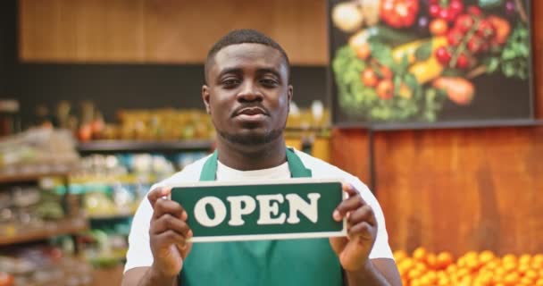 Pohledný mladík stojí za stánkem a drží prkno v obchodě. Afro-americký obchodník v zástěře ukazuje ceduli OPEN a dívá se na kameru v supermarketu. Obchod, obchodní koncept. — Stock video