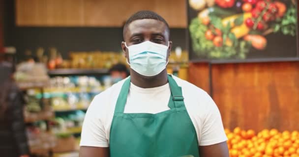 Incolpare il venditore maschile in maschera protettiva medica che lavora nel negozio di alimentari. Il giovane afro-americano guarda la telecamera e mostra il cartello OPEN. Misure di sicurezza, coronavirus, concetto covid-19. — Video Stock