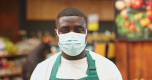 Portrait of good-looking adult man in medical protective mask standing in superarket. Afro-American salesman guy looking at camera in grocery store. Coronavirus, covid-19, pandemic, safety concept. — Stok video