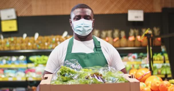 Привлекательный работник мужчина в медицинской защитной маске работает в продуктовом магазине. Молодой афро-американец держит коробку с салатом и смотрит в камеру в супермаркете. Коронавирус, концепция здоровья. — стоковое видео