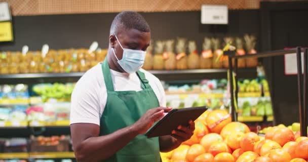 Bello il conteggio dei dipendenti maschi e la segnalazione dei dati su tablet in negozio di alimentari. Un giovane afro-americano con una maschera medica, che lavora al supermercato. Marketing, lavoro, concetto di coronavirus. — Video Stock