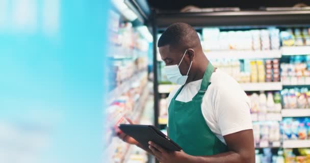 Jonge Afro-Amerikaanse man in masker maken verslag over zuivelproducten in de buurt refregeration sectie. Aantrekkelijke mannelijke werknemer tellen spullen op planken, typen op tablet in de supermarkt. Tellen, zakendoen. — Stockvideo