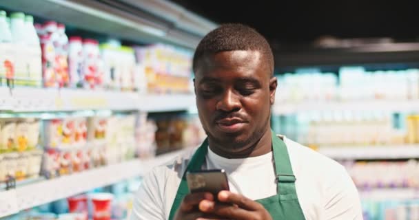 Young Afro-American guy in apron using smartphone and looking at dairy prodcuts shelves. Handsome salesman scrolling feed, texting and smiling in supermarket. Social media, commerce concept. — Stok video