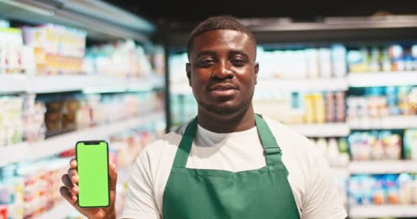 Der erwachsene männliche Geschäftsinhaber zeigt einen grünen Bildschirm und blickt in die Kamera in der Milchabteilung. Charmanter Mann hält Smartphone mit Chroma-Schlüssel in der Hand und lächelt im Supermarkt. Handel, Technologie. — Stockvideo